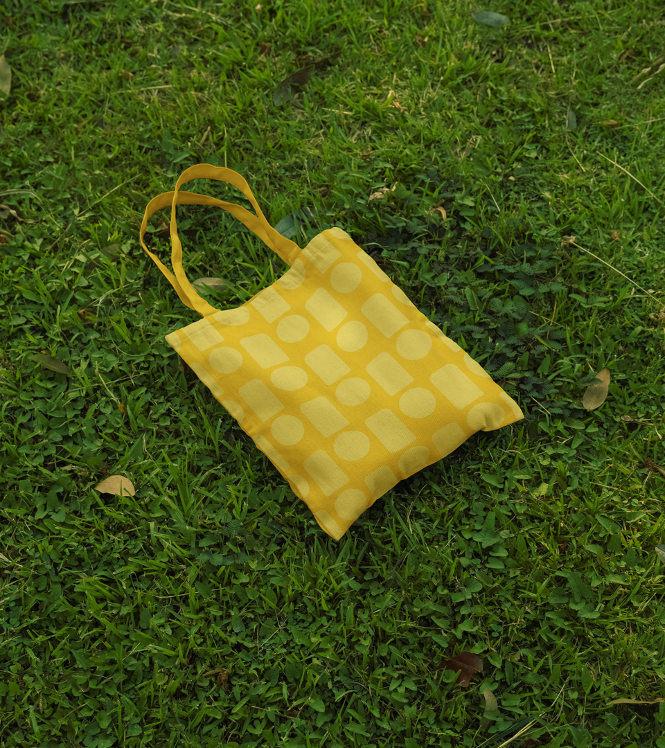 Mockup of a yellow tote bag with a Pinni-branded pattern, laid on grass.